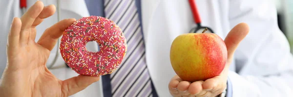 Doctor in office holding in hand pink doughnut vs red apple — Stock Photo, Image