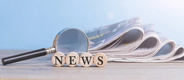 Writes news on wooden cube with newspaper stack and magnifying g — Stock Photo, Image