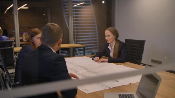 I det moderna kontoret tre anställda vid bordet ser ritpapper och prata. — Stockvideo