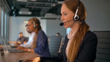 Anlaşma ofisinde çalışan merkezi işçilerin ara. İlk plan müşteriyle konuşurken kulaklık yakışıklı genç bayan. Güzel manzaralı modern iş oda. Ağır çekim yakın çekim.