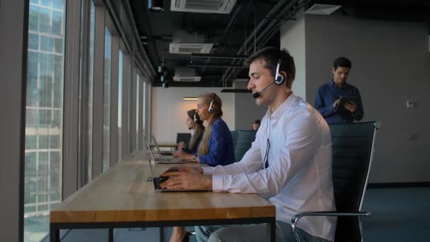 Man in het shirt een hoofdtelefoon met laptop gebruikt terwijl u werkt in een call center office — Stockvideo