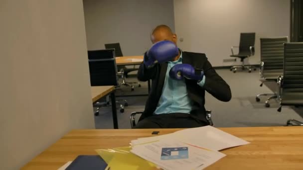 Sporty fighty leader in business concept. Afro-American businessman sitting with boxing gloves in office — Stock Video