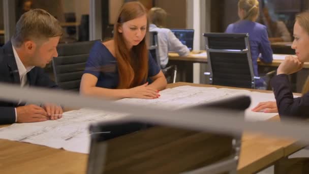 In the modern office three staff at the table look the drawing paper and talking — Stock Video