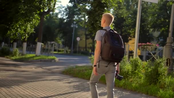 Back-side weergave van student fotograaf met een zwarte rugzak loopt langs een Dorpsstraat — Stockvideo