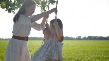 Beyaz otantik elbiseli nazik mutlu anne sahada salıncak onu güzel kızları sallar