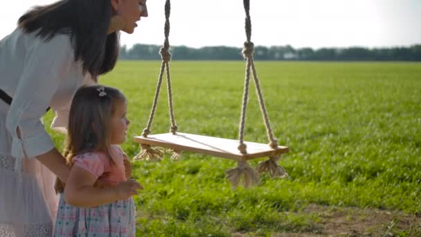 Красива мати в білій сукні допомагає доньці сидіти на дерев'яній підвісній мотузці . — стокове відео