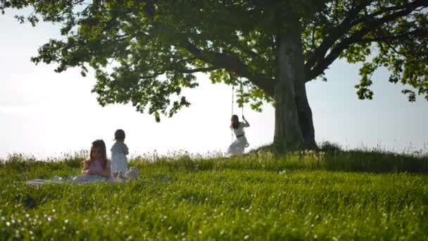 Mooie vrouw in een rustieke witte jurk swingend op een schommel, haar dochters in het veld. — Stockvideo