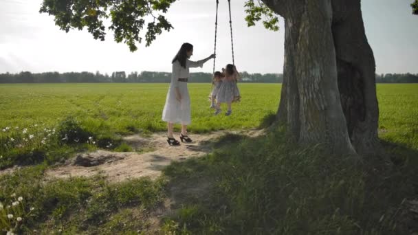 Молодая мать нежно трясет своими двумя дочерьми 1 и 3 лет на качелях — стоковое видео