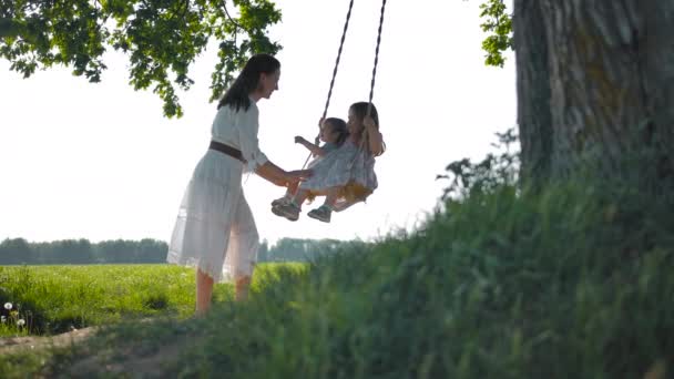 Mamá balancea a sus dos hijas felices en un columpio de cuerda en medio de un campo verde — Vídeos de Stock