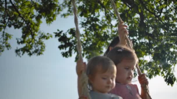 Mamma skakar två små systrar på en gunga med rep handtag — Stockvideo