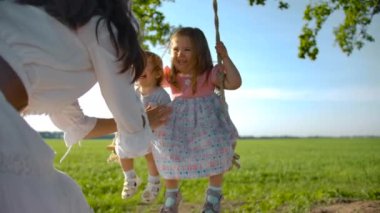 Beyaz elbiseli mutlu kadın kızlarını salıncakta sallıyor.