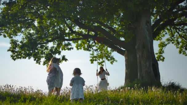 Två döttrar går längs gröna fältet till sin mor, svänga på ett rep Swing knuten till gamla träd. — Stockvideo