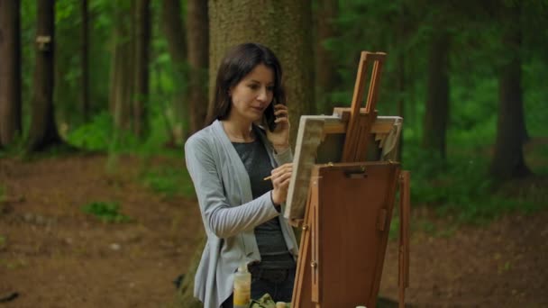 Chica artista pintando un cuadro en el parque y hablando por teléfono — Vídeos de Stock