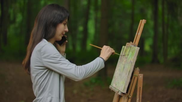 Vista lateral media de la artista chica pintando un paisaje en el parque y hablando por teléfono — Vídeos de Stock