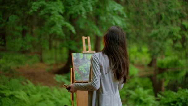 Rear view of a young artist female painting a landscape and wipes the brush in a summer forest — 图库视频影像