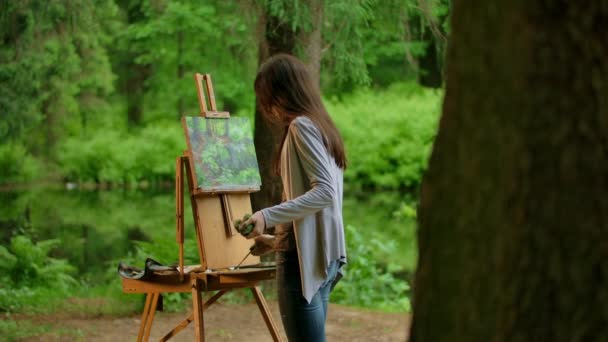 Vista trasera del medio de una joven artista pintando un paisaje en un bosque de verano — Vídeos de Stock