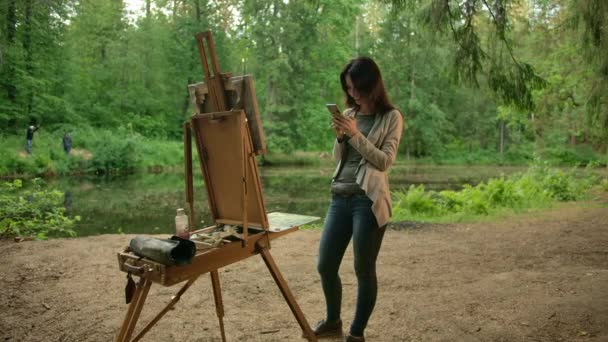 Девушка-художник, рядом с мольбертом и палитрой, общается со своими друзьями в смартфоне . — стоковое видео