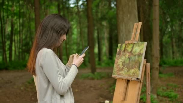 Девушка-художник закончила художественные работы над своей картиной и чата в своем смартфоне — стоковое видео
