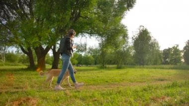 Parkta köpeğiyle yürüyen kulaklıklı bir kızın paralel görüntüsü..