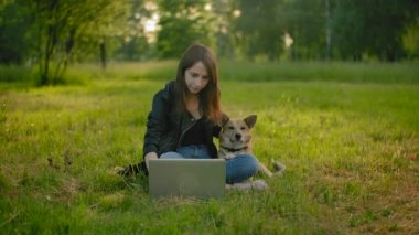 Parkta dizüstü bilgisayarın yanında beyaz bir kadın köpeğini okşuyor..