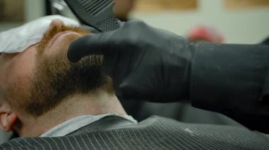 Gözlerinde havluyla genç bir adamın sakalını kesiyor..