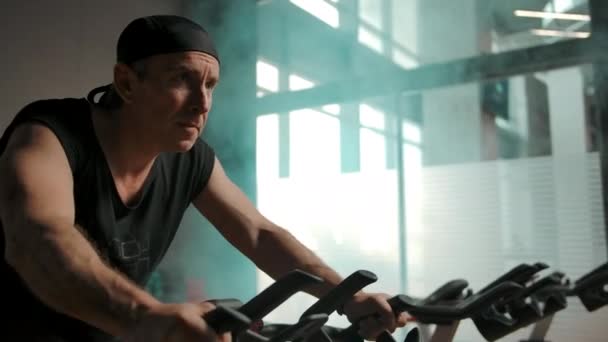 Homme adulte en T-shirt noir et bandana trains sur vélo d'arrêt dans une chambre bizutée — Video