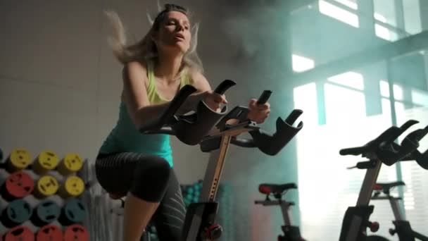 Tozluklu ve tişörtlü güzel bir kız bisiklet eğitimi aletinde pedal çeviriyor. — Stok video