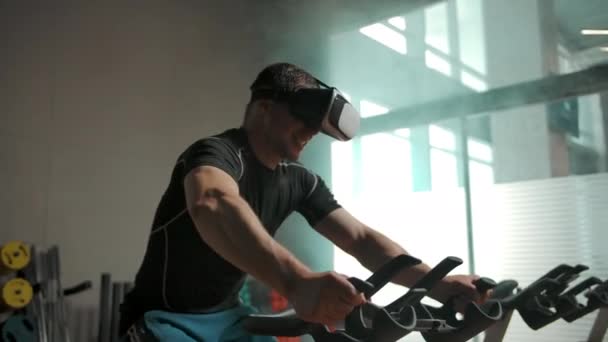 Een man doet een zware oefening op een fietssimulator in een VR helm — Stockvideo