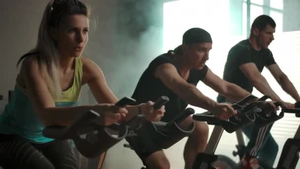 Les sportifs entraînent les muscles des jambes sur le vélo d'appartement dans la salle de gym en rangée — Video