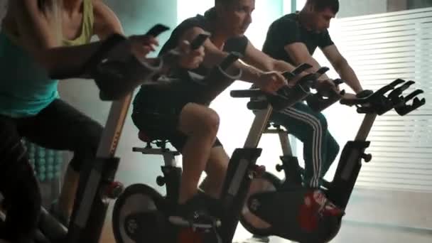 Sportmen entraînement des muscles des jambes sur les vélos d'exercice dans la salle de gym dans une rangée. — Video