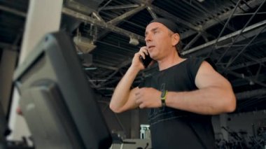Erkek sporcu koşu bandında koşarken cep telefonuyla konuşuyor.