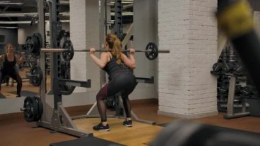 Spor salonunun içinde halterle çömelme hareketi yapan seksi kadın antrenörün arka görüntüsü