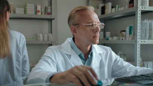 En mogen farmaceut med glasögon sittande vid en dator på ett apotek. — Stockvideo
