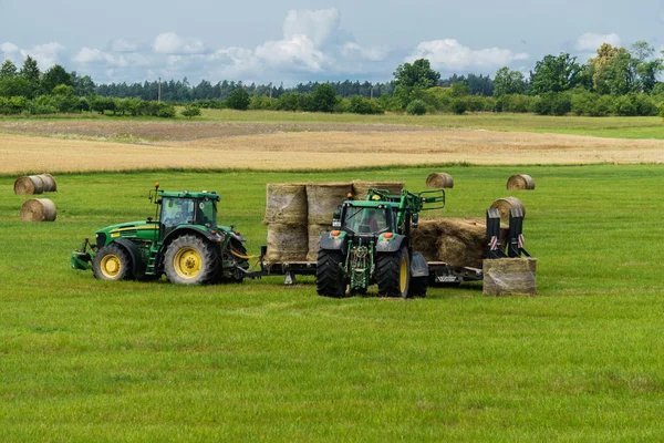 A tractor loads bales of hay onto a trailer from another tractor.