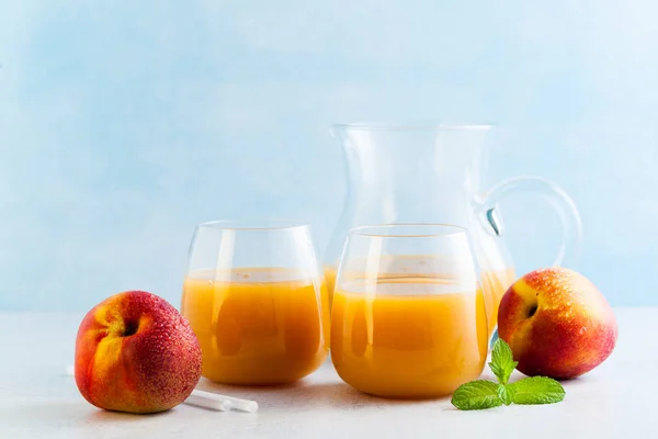 peach juice in glasses and a decanter and fresh peaches with leaves on a table. blue background. copy space