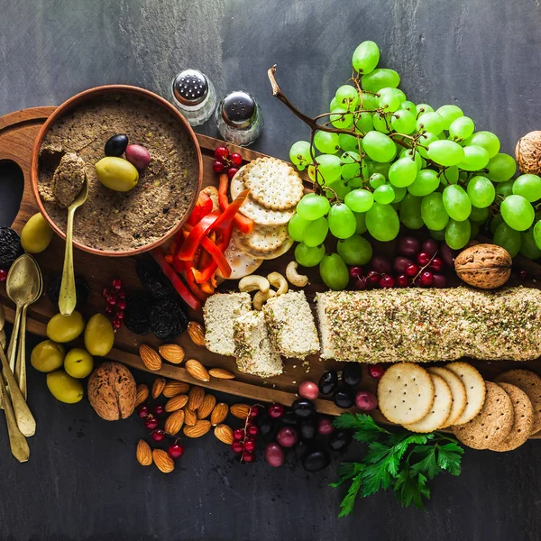 Wooden serving board with vegan snacks. Pate of olives and cheese from cashew nuts with herbs. Healthy appetizer with grapes, nuts, and dried fruits on a black slate table.