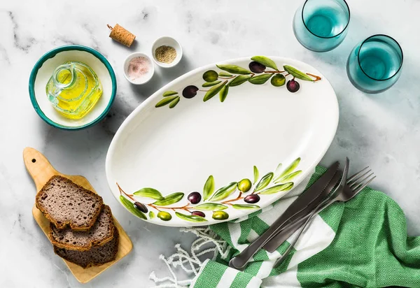 Empty handmade oval dish with olive tree branches on a white marble table. olive oil and cutlery. Mediterranean table setting