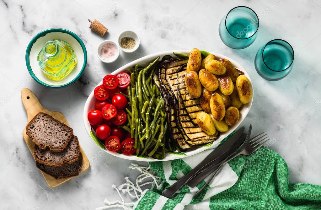 a side dish of vegetables on the holiday table. healthy food for the whole family or dinner at a restaurant on a white marble table. baked potatoes, grilled eggplants, cherry tomato salad and steamed green beans with garlic sauce.