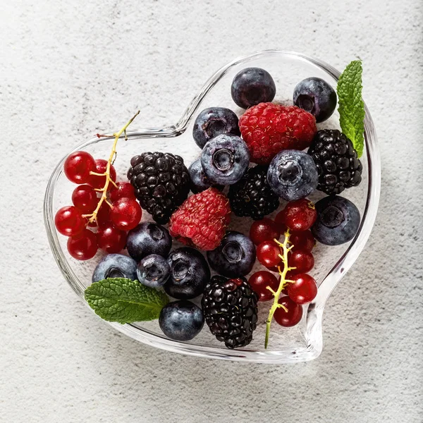 Bagas maduras frescas em uma tigela em forma de coração de vidro em uma pedra branca — Fotografia de Stock