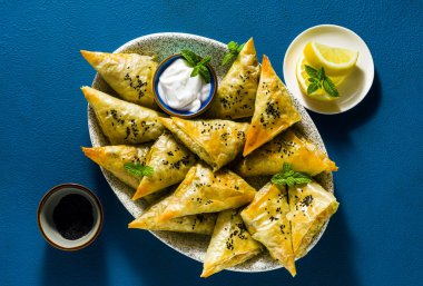 Baharatlı patates ve sebzeli Hint samosası. Mavi arka planda yoğurt, nane ve limonla servis ediliyor.