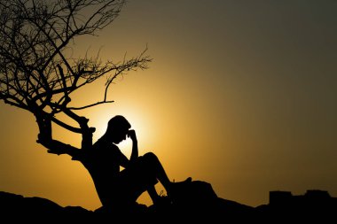 Gün batımında ağacın yanında oturan bir adamın silueti.