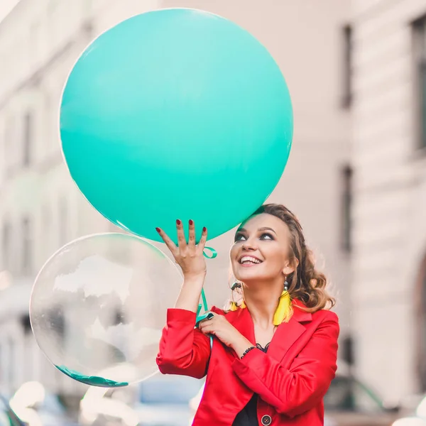 一个美丽的明亮的女孩在城市街道上的肖像绿色气球的微笑和摆在红色西装 — 图库照片