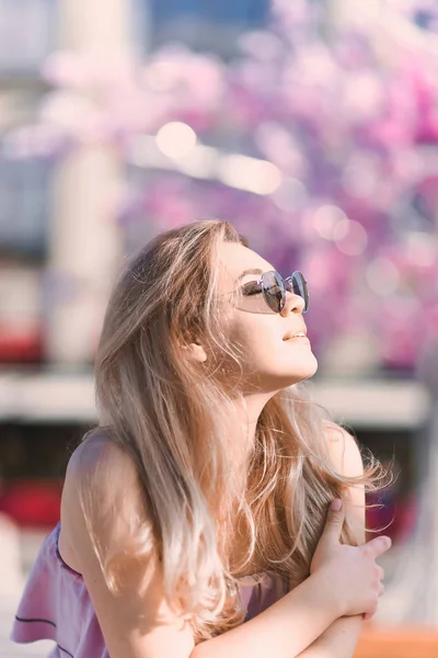 肖像美丽的年轻金发女孩在太阳镜与肿胀的嘴唇和性感的女性身体摆在街上与粉红色的花朵在阳光明媚的一天背景 — 图库照片
