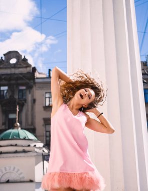 yakın çekim yaşam tarzı bir sokakta gülümseyen güneşli bir günde bir pembe tüy elbiseli kıvırcık saçlı Genç güzel seksi kız portresi