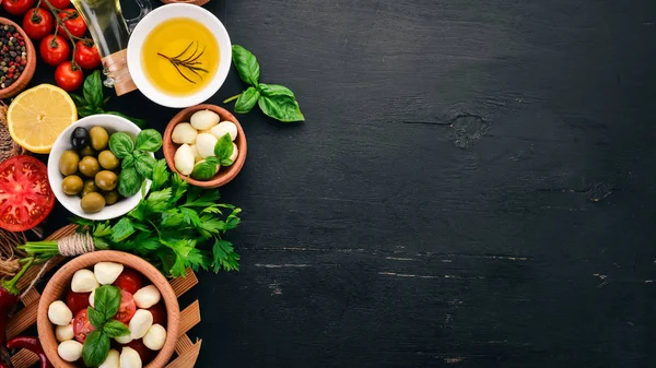 Ingredients for Italian caprese salad. Mozzarella cheese, cherry tomatoes, basil leaves, olives, oil, pepper. On a black wooden background. Free space for text.