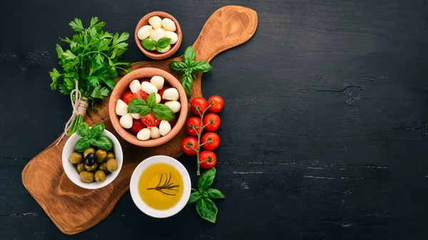 Ingredients for Italian caprese salad. Mozzarella cheese, cherry tomatoes, basil leaves, olives, oil, pepper. On a black wooden background. Free space for text.