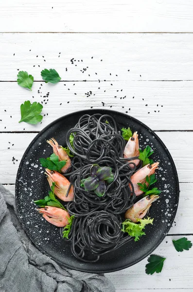 Black pasta with shrimps on a black plate. Top view. On a white wooden background. Free copy space.