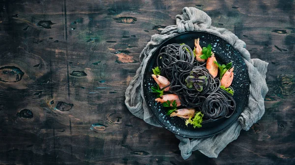 Black pasta with shrimps on a black plate. Top view. On a white wooden background. Free copy space.