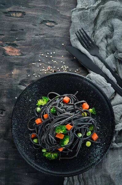 Black pasta with vegetables on a black plate. Top view. On a white wooden background. Free copy space