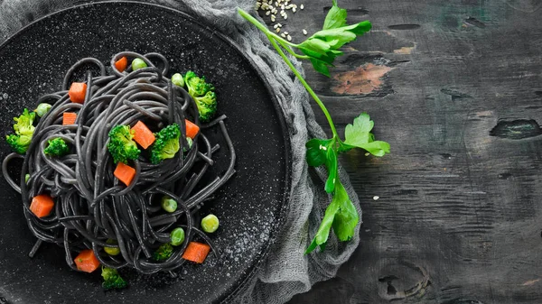 Black pasta with vegetables on a black plate. Top view. On a white wooden background. Free copy space